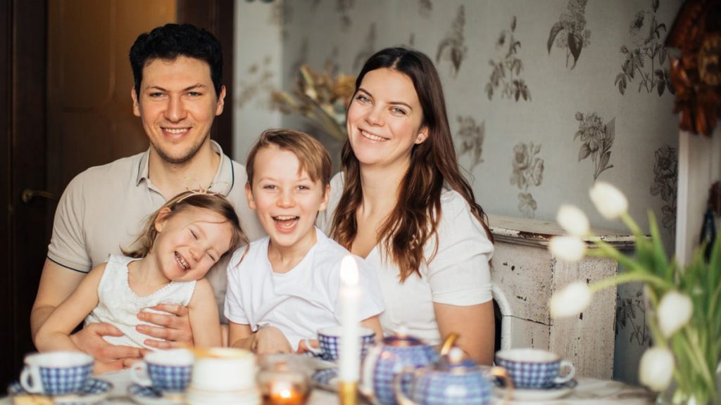 Positive family in casual clothes enjoying tea with sweets sitting at table set for breakfast with candle and laughing while looking at camera